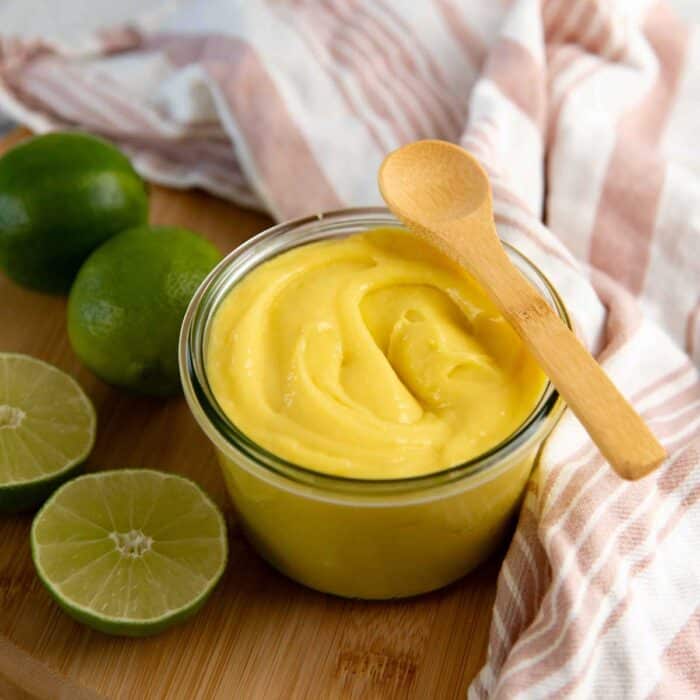Glass jar of creamy lime curd with a small wooden spoon on top, and fresh limes nearby.