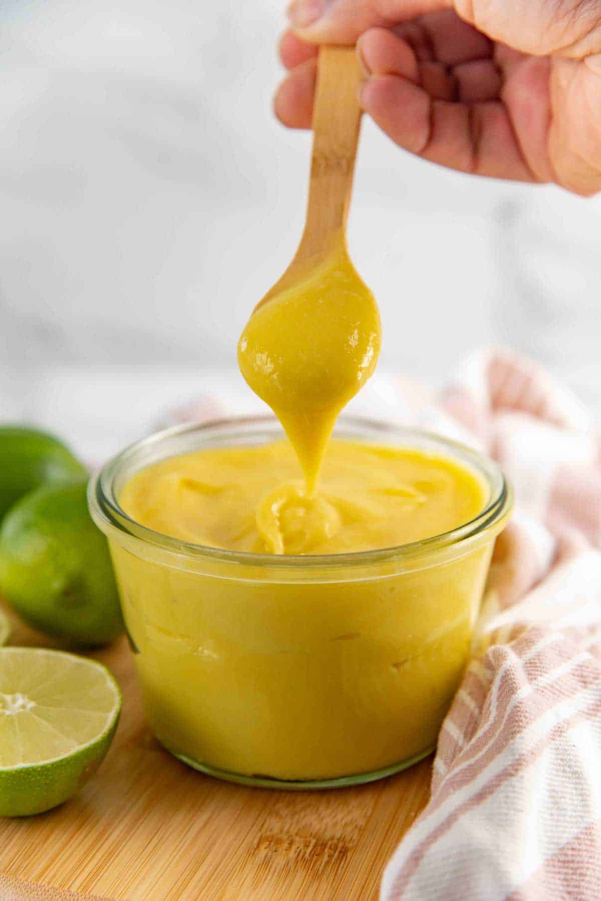 A hand holds a wooden spoon dripping thick yellow lime curd into a glass jar full of lime curd.