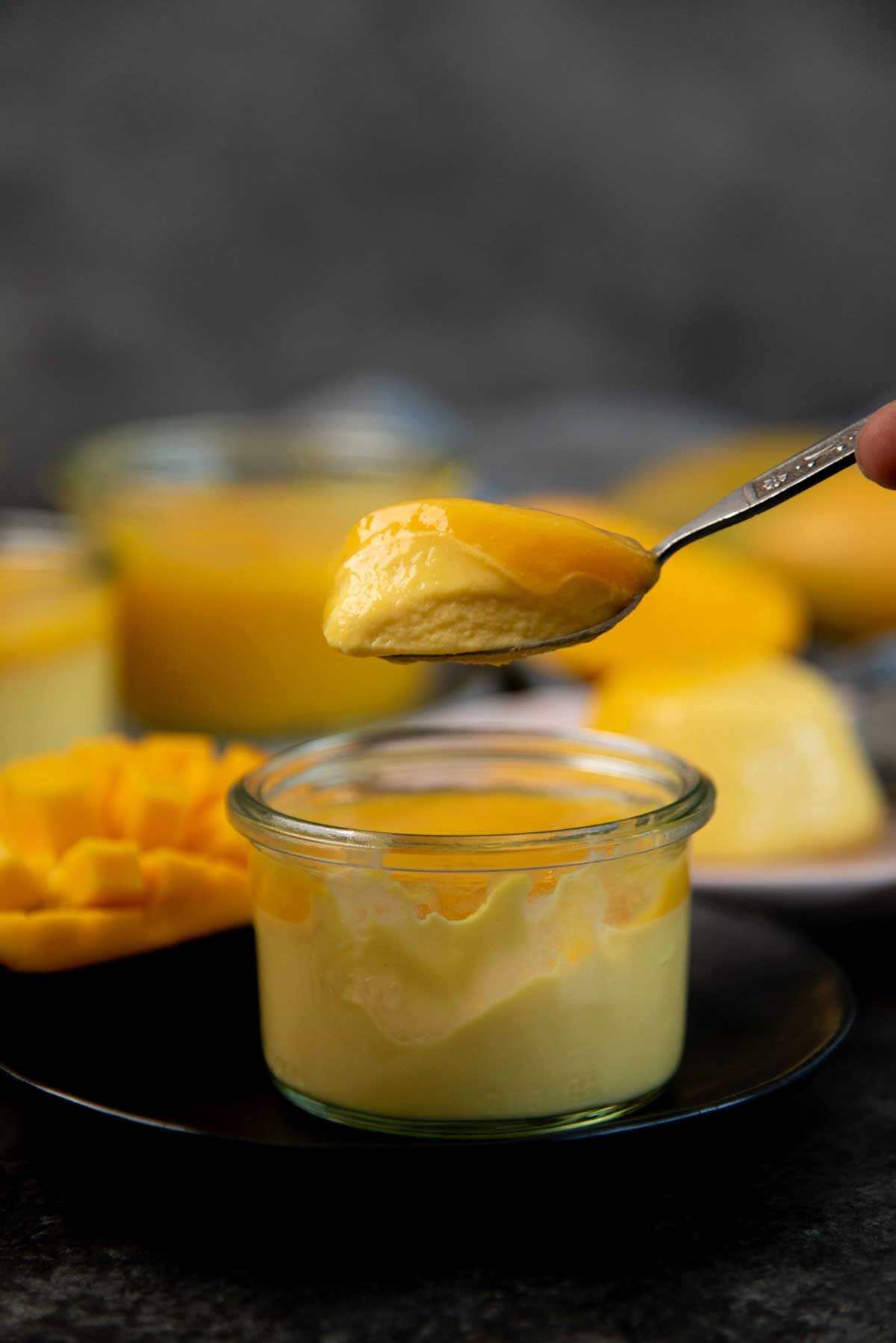 A spoon holds creamy yellow mango panna cotta over a glass jar, with a sliced mango in the background.