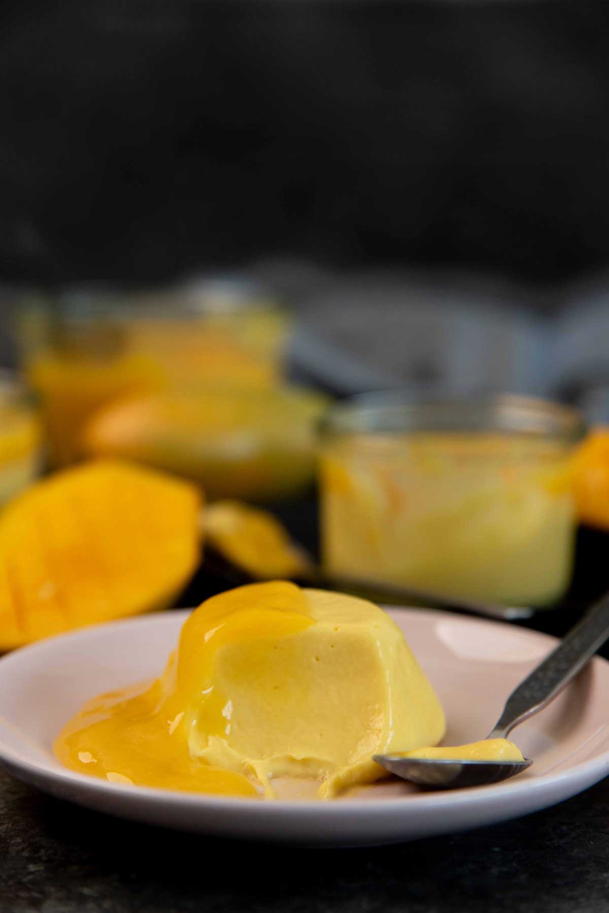 A creamy mango dessert served on a pink plate with mango gel sauce dripping, accompanied by a spoon.