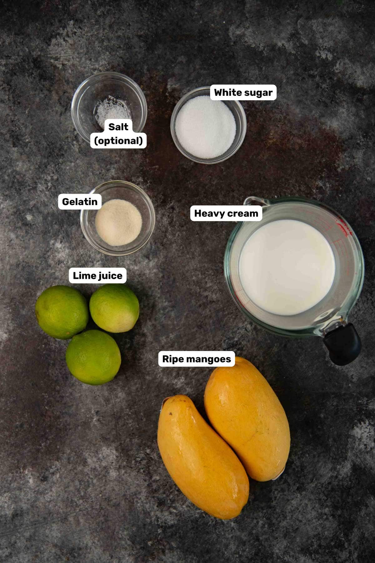 Labeled ingredients to make mango panna cotta on a dark surface.