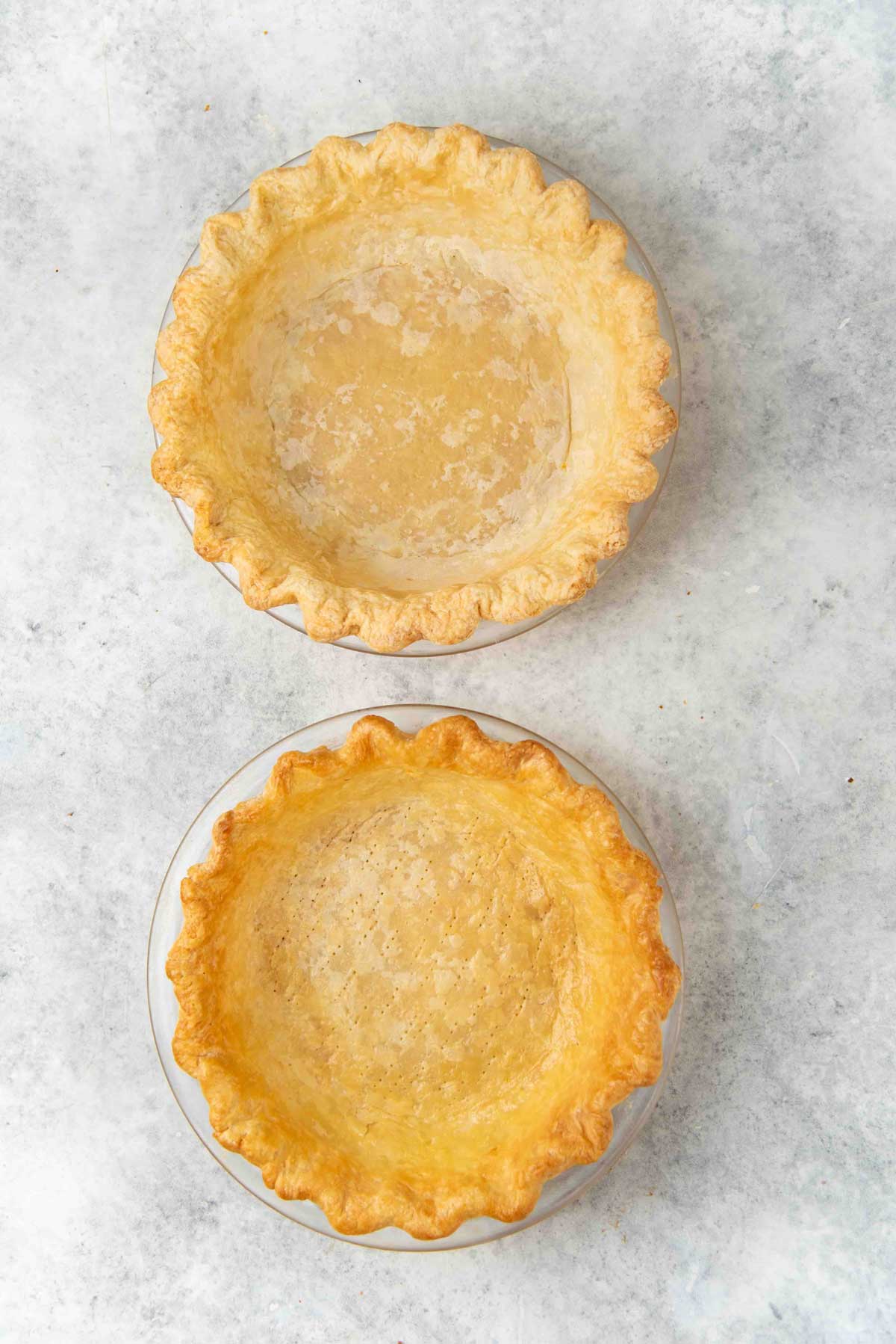 Two flaky pie crusts in round baking dishes. The one on top is par baked, the one below is fully blind baked and has a deeper golden brown color.