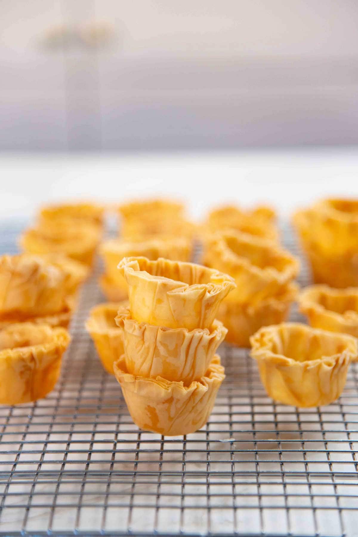 Baked mini phyllo cups stacked on a cooling rack, surrounded by more phyllo cups.