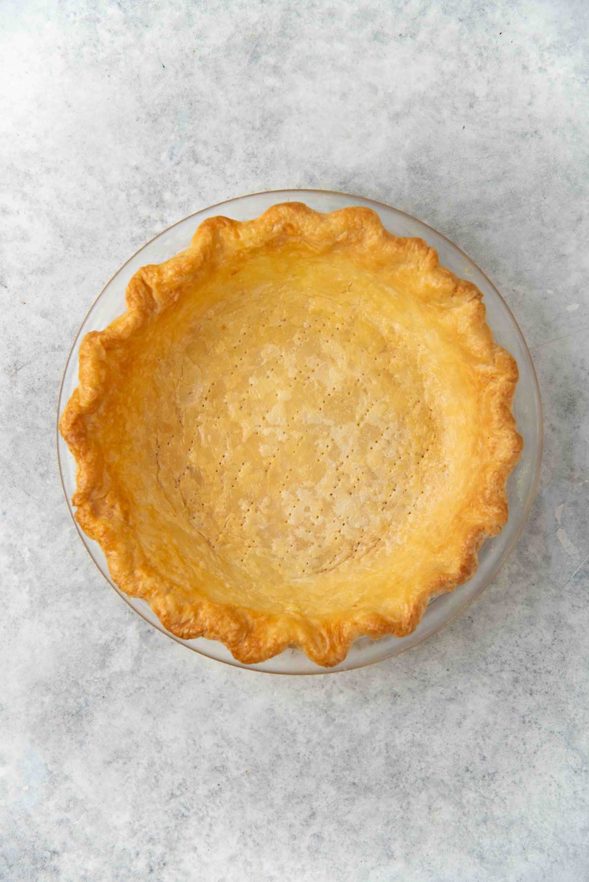 Golden brown, flaky blind baked pie crust in a round glass pie dish, with a crimped edge against a light background.