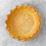 Square image of blind baked pie crust in a round glass pie dish, with a crimped edge against a light background.