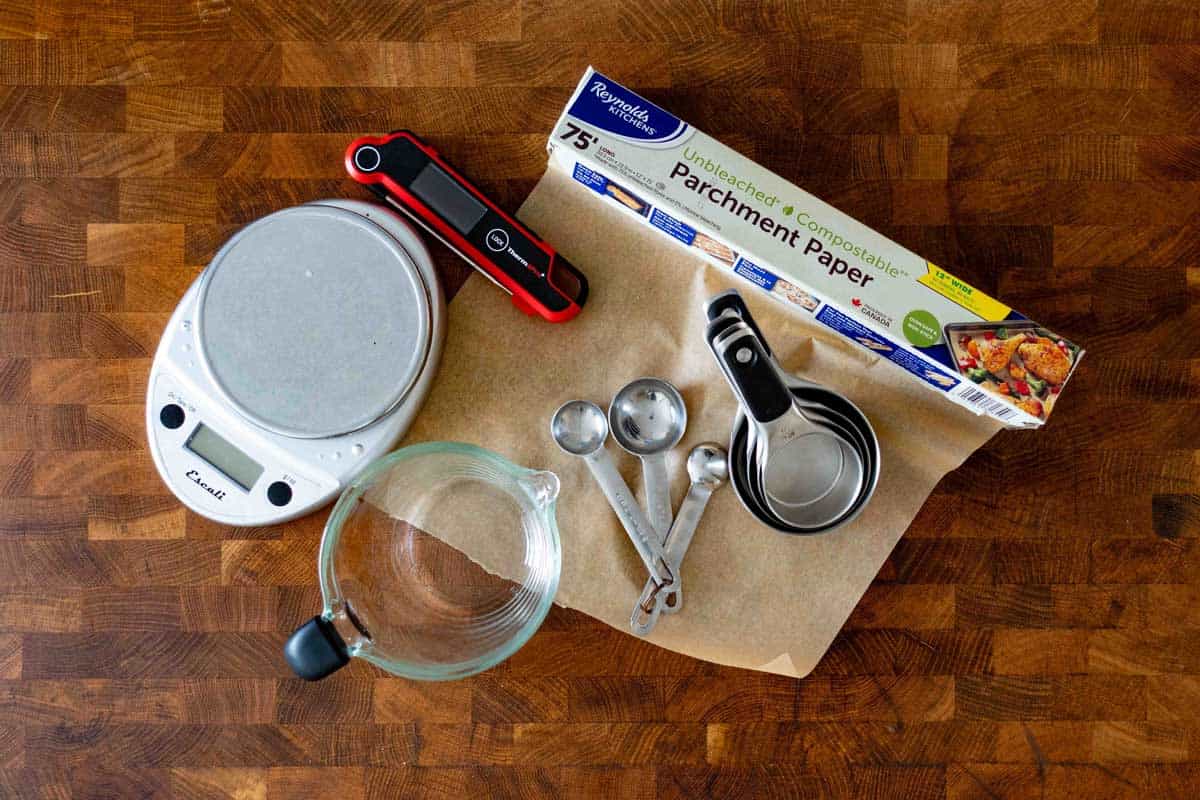 Baking tools on a wooden table including digital scale, red thermometer, parchment paper, glass bowl, metal measuring cups and spoons.