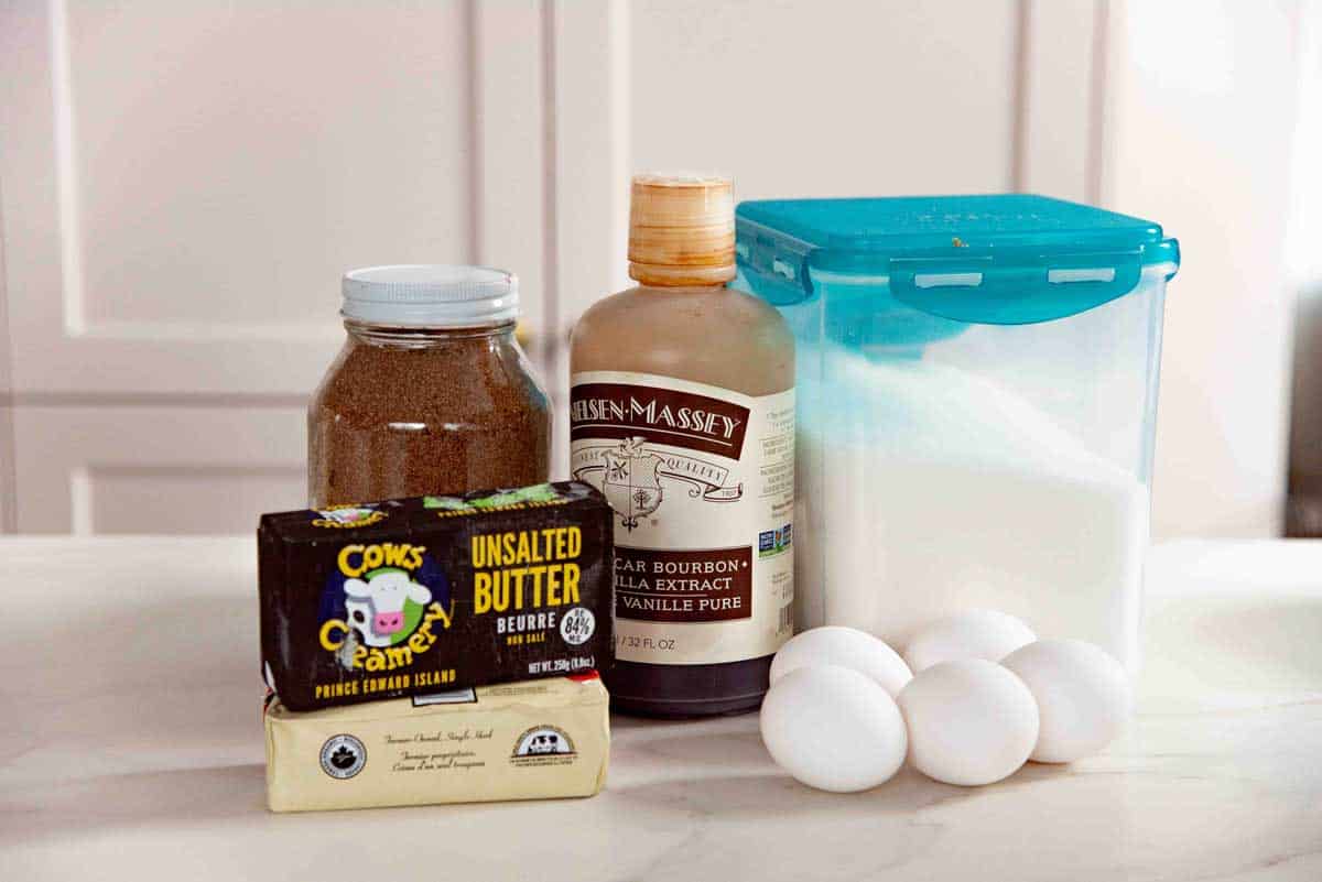 Basic pantry ingredients for baking including white sugar, eggs, butter, vanilla extract, and brown sugar on a white surface.