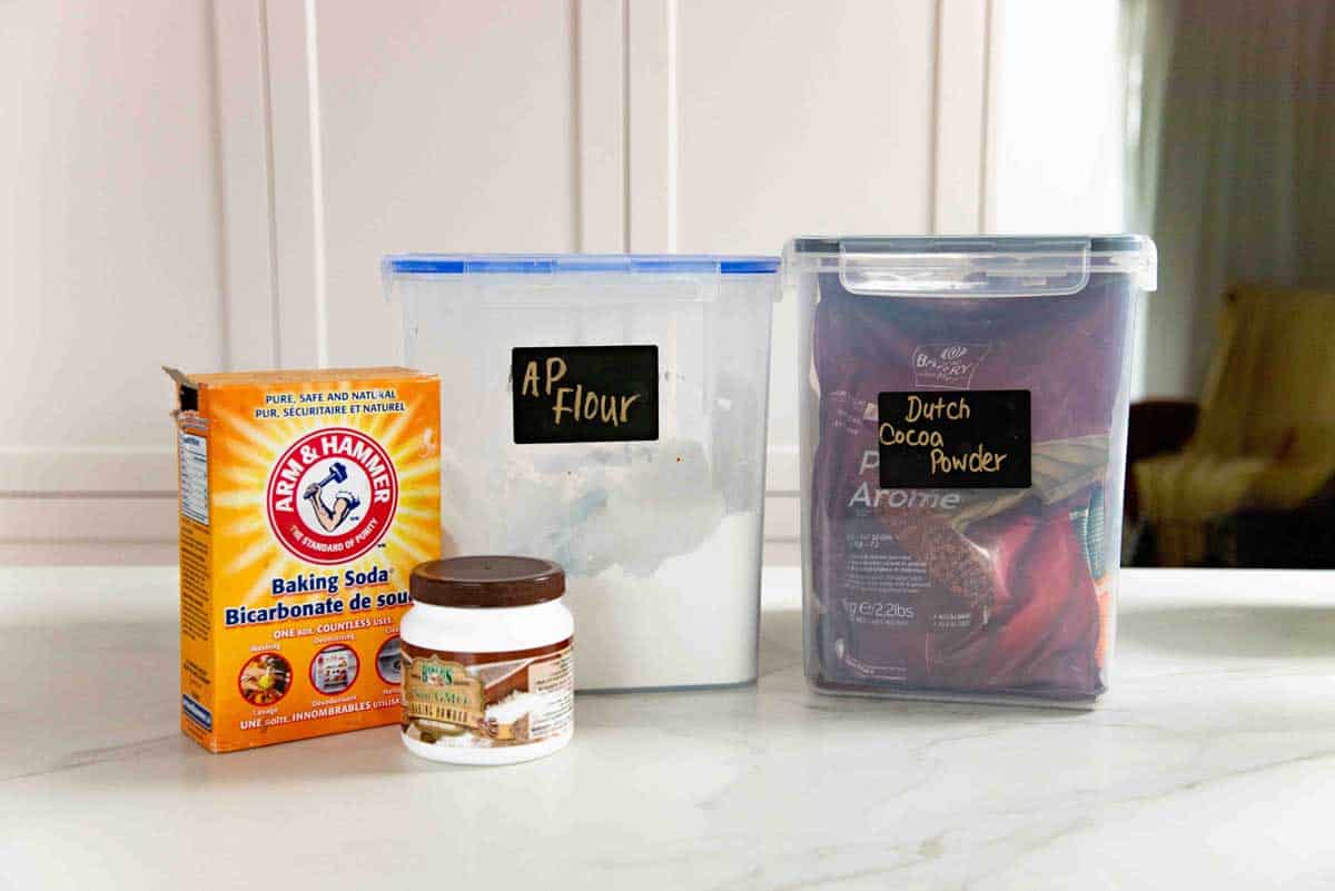 Baking soda, baking powder, all purpose flour and cocoa powder in separate containers on a white countertop.