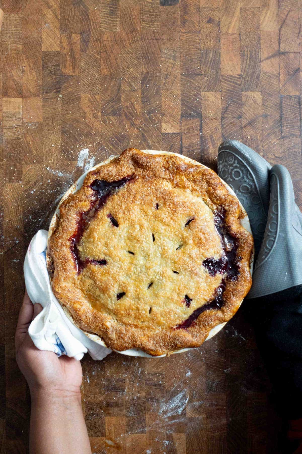A golden round pie with a flaky double crust and blueberry filling, held by a hand with a white cloth on a wood surface.