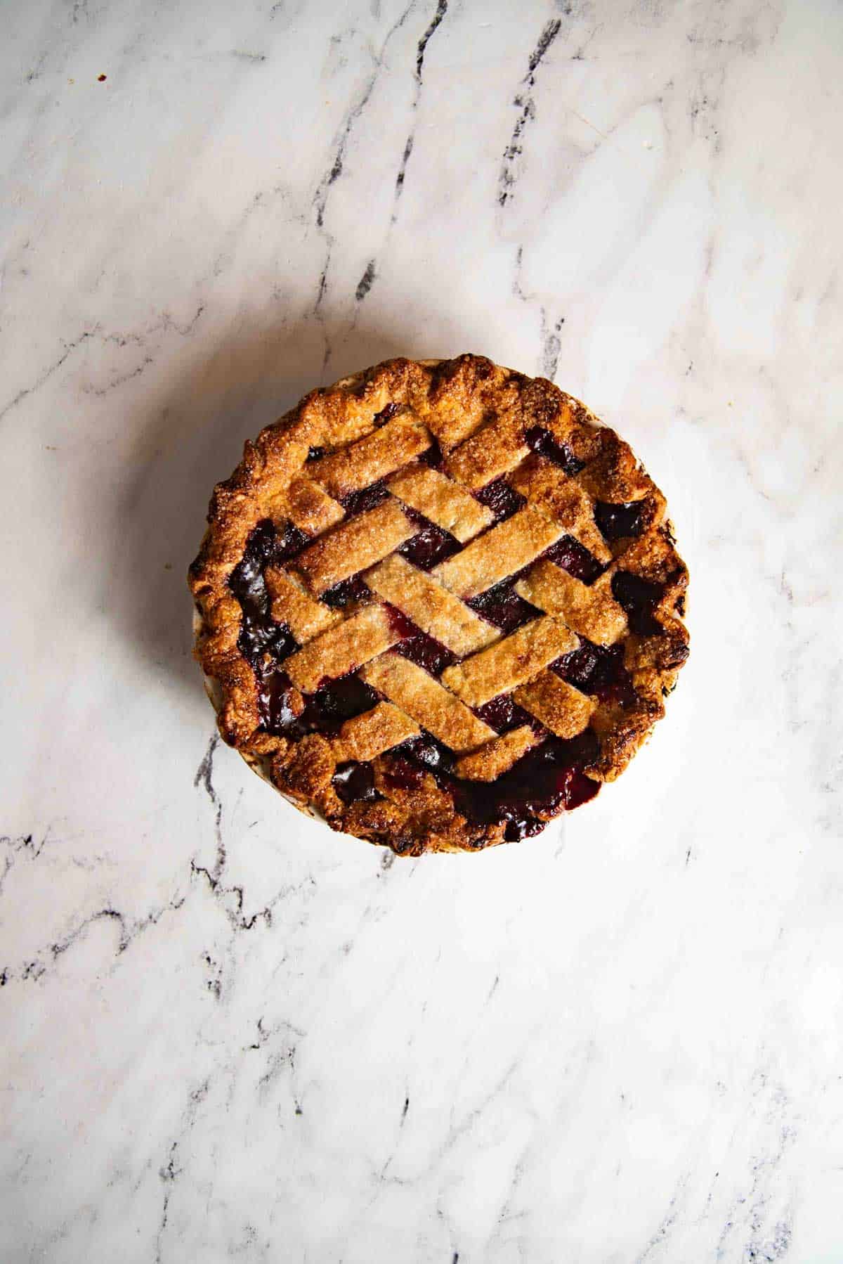 Freshly baked round pie with a golden, flaky lattice pie crust against a white surface. The filling can be seen through the gaps in the lattice crust.
