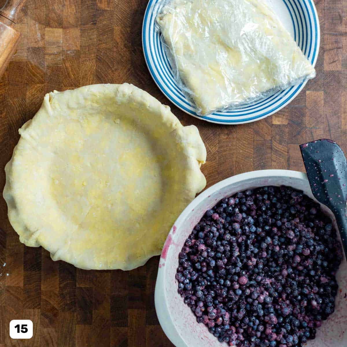 A wooden table with a pie crust in a dish, dough portion wrapped in plastic wrap, and a bowl of wild blueberry filling.