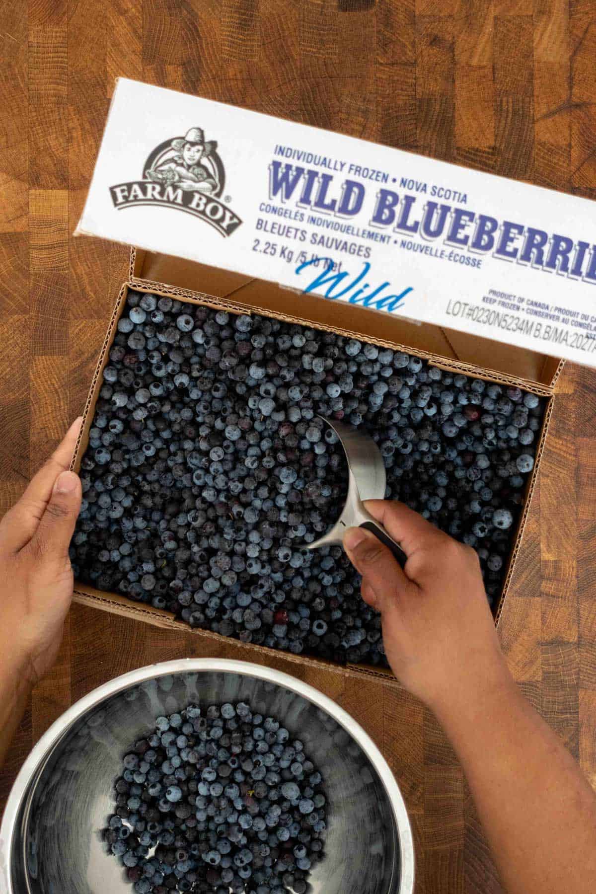 Hands scoop frozen wild blueberries from a box into a metal bowl on a wooden surface.