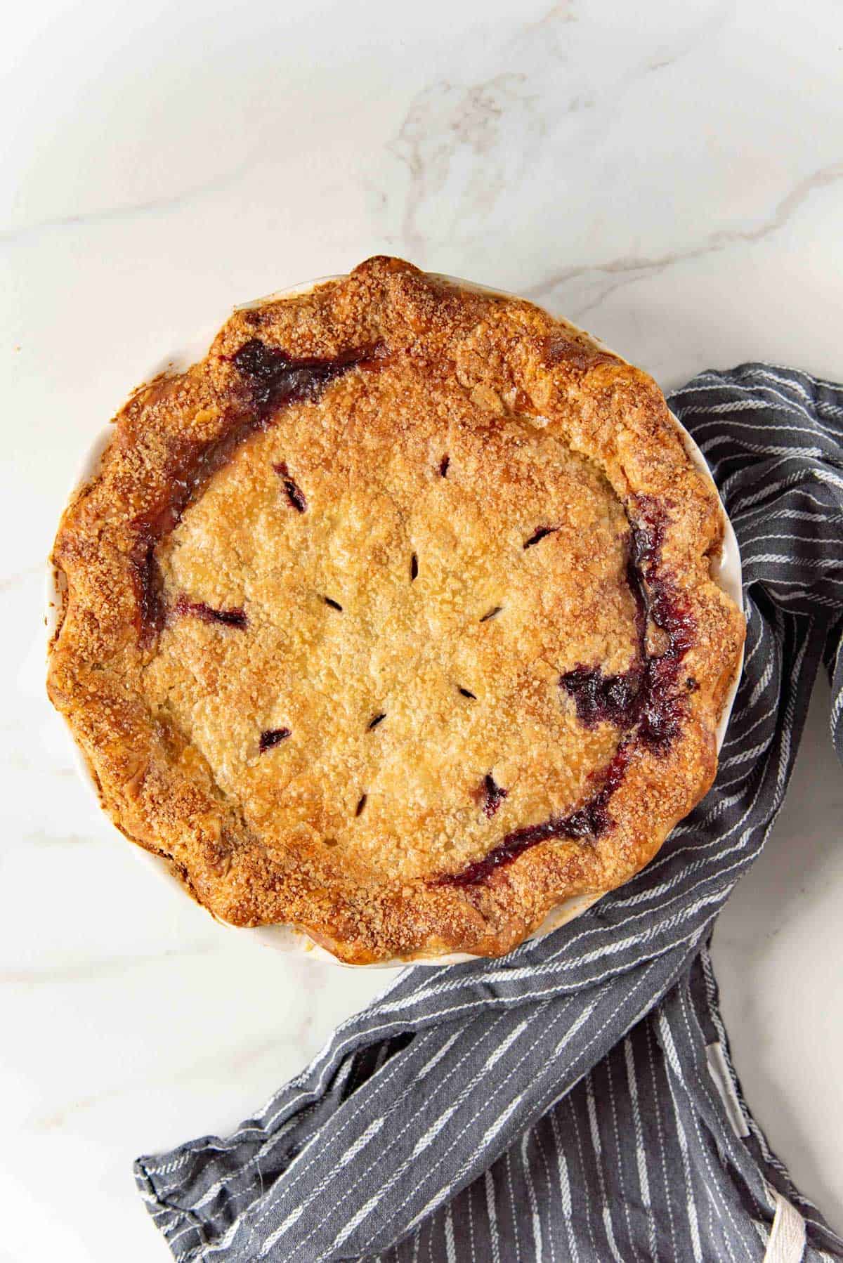 Overhead image of a blueberry pie with a flaky golden solid top crust on a light colored countertop.