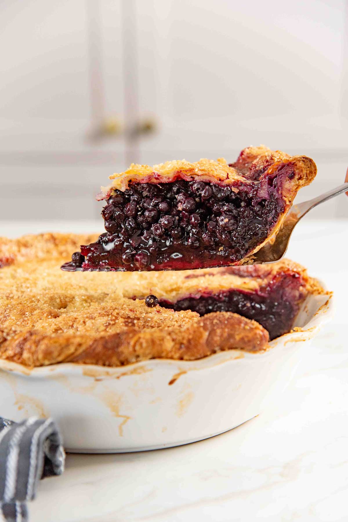 A slice of blueberry pie with a golden, flaky crust is being lifted from a pie dish with a serving spoon. The filling is rich and juicy.
