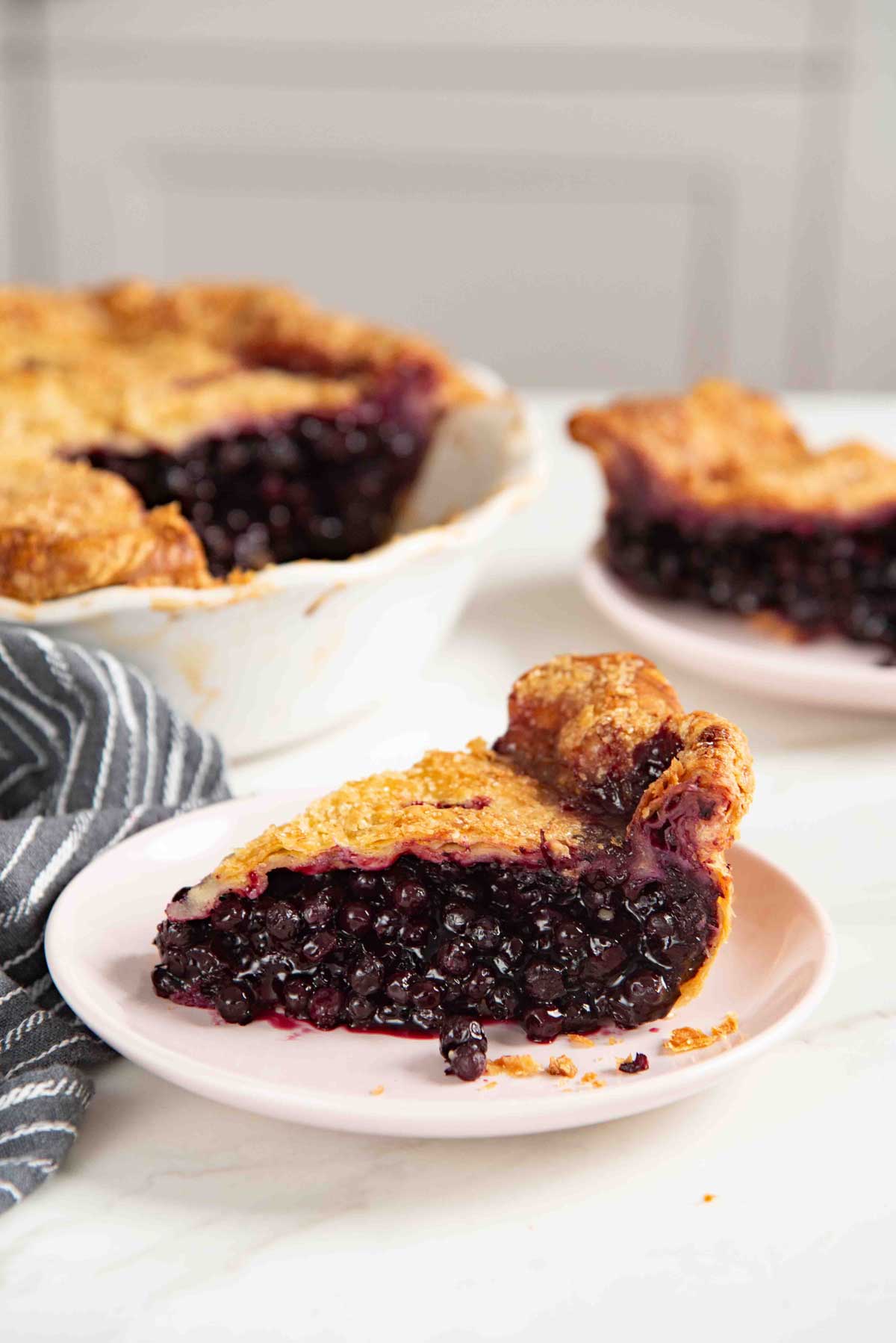 Slice of blueberry pie with a flaky golden crust and a juicy wild blueberry filling served on a small pink plate.