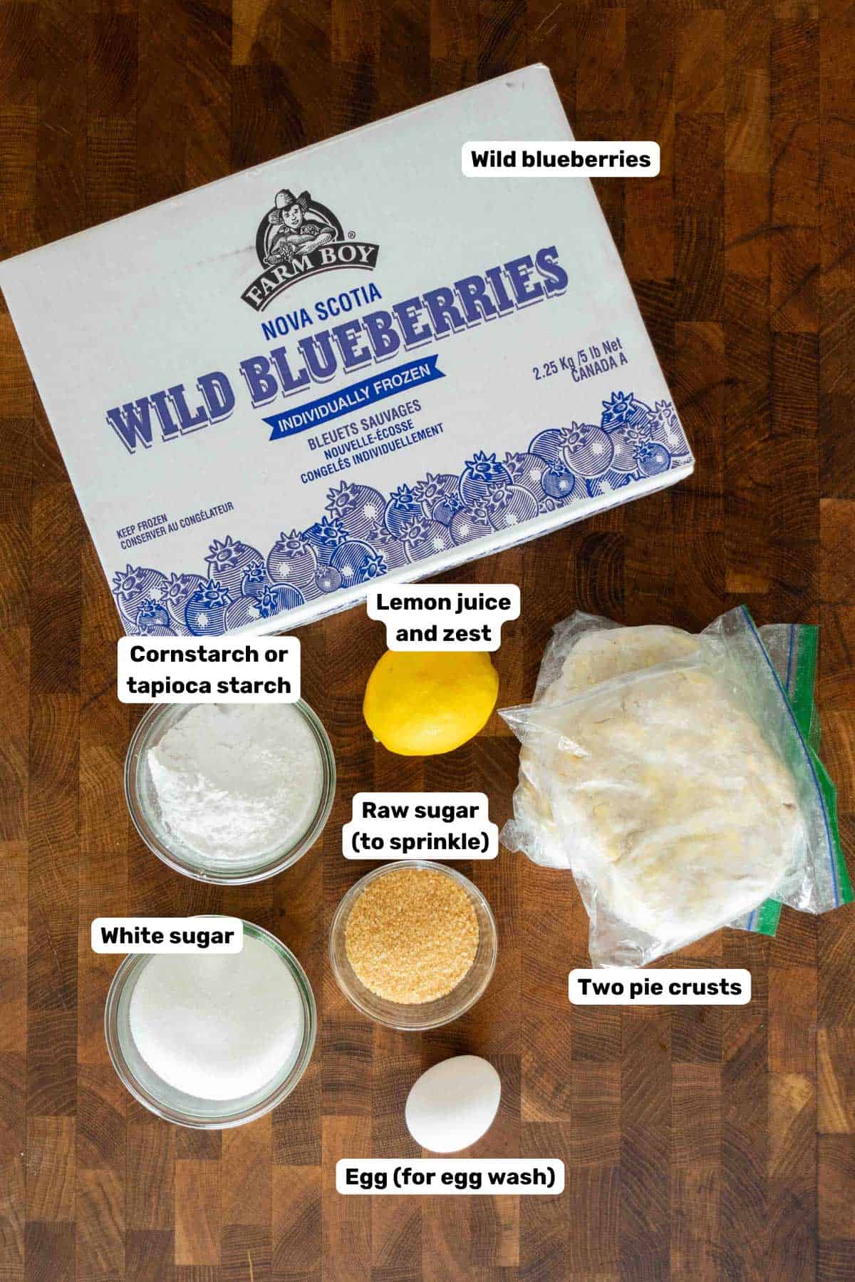 Labeled ingredients to make wild blueberry pie on a wooden surface.