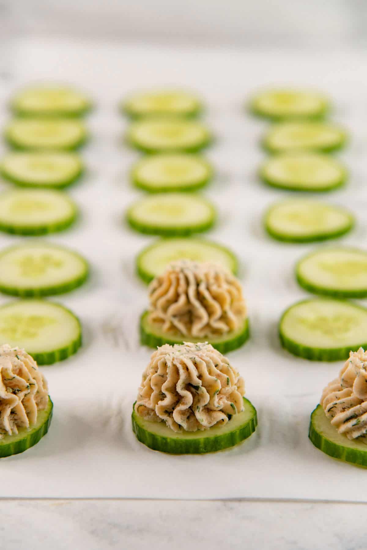 Rows of round cucumber slices on a parchment paper with creamy smoked salmon mousse piped onto several of them.