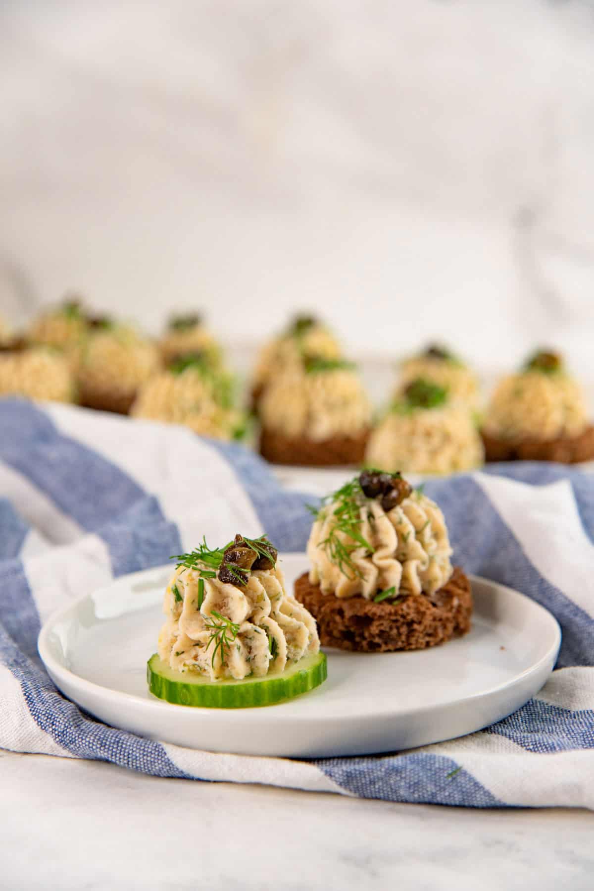 A white plate with a cucumber slice and a rye bread slice topped with salmon mousse, chopped herbs and capers.