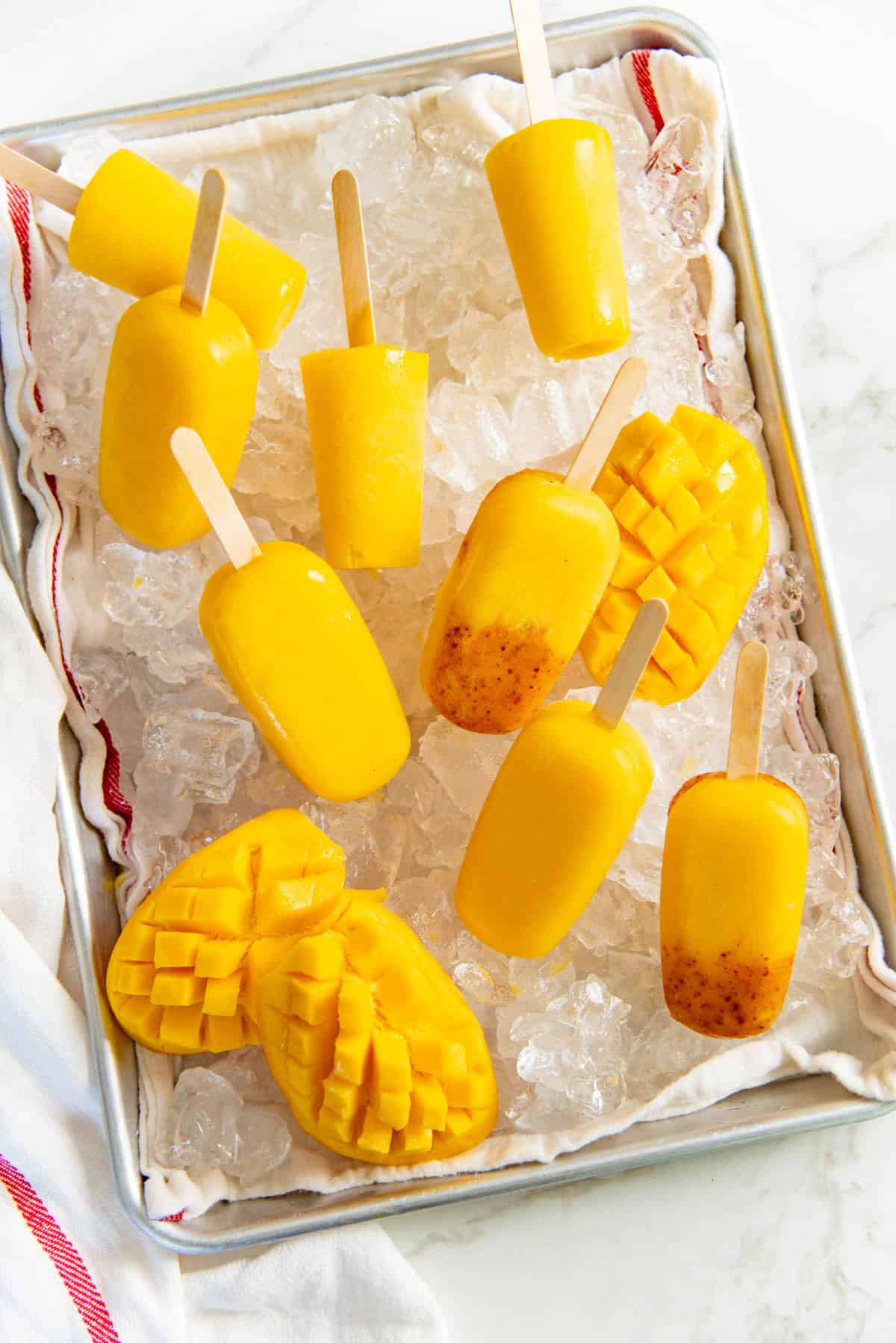 Overhead view of mango popsicles, mango tajin popsicles, and freshly cut mangoes on a bed of ice in a sheet pan.