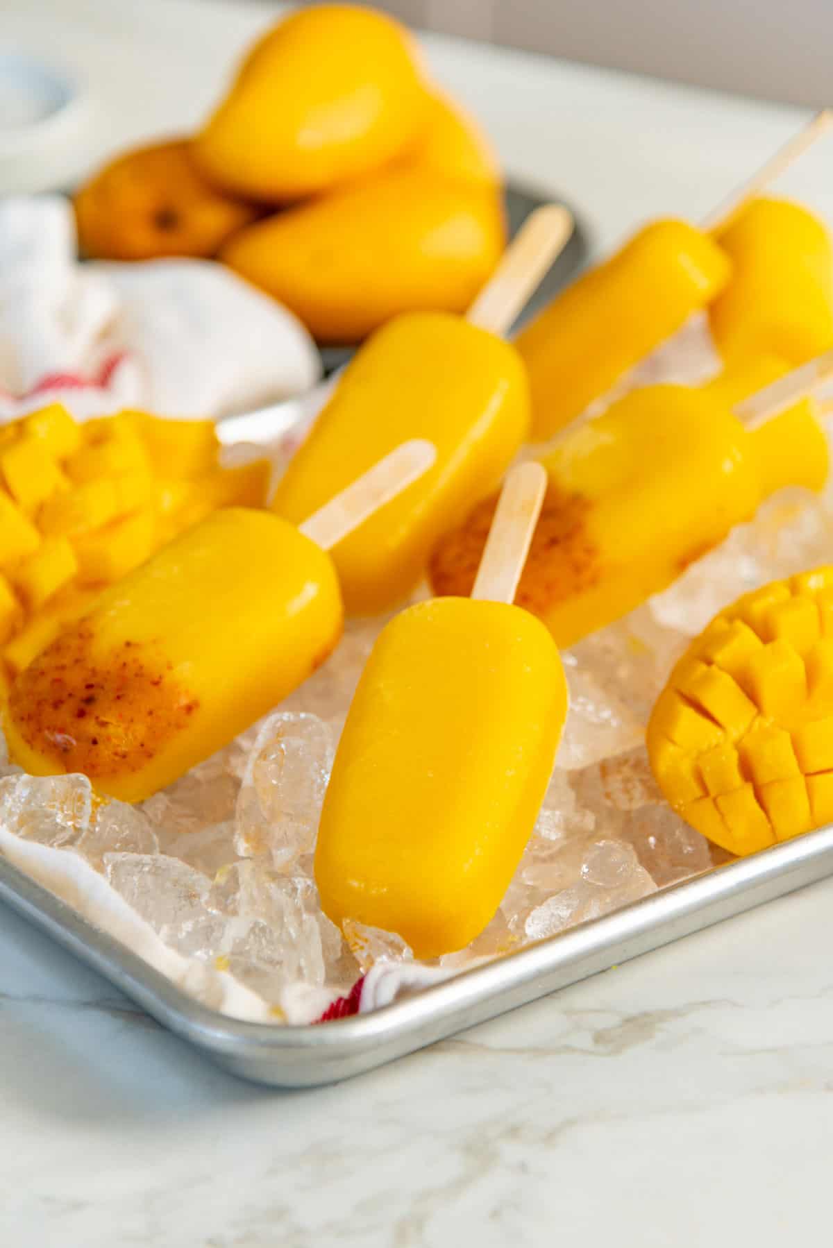 Yellow fruit popsicles and freshly sliced and cut mangoes placed on top of ice in a baking pan.