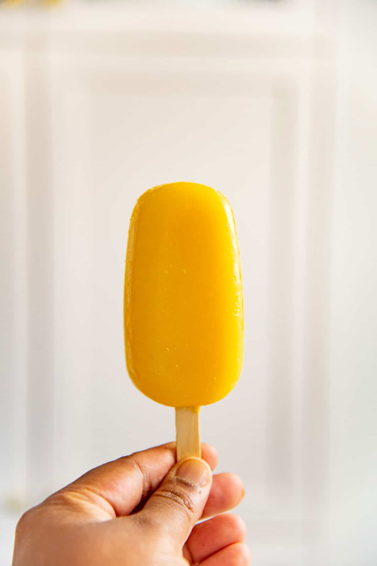A hand holding up a bright yellow mango popsicles against a white background.