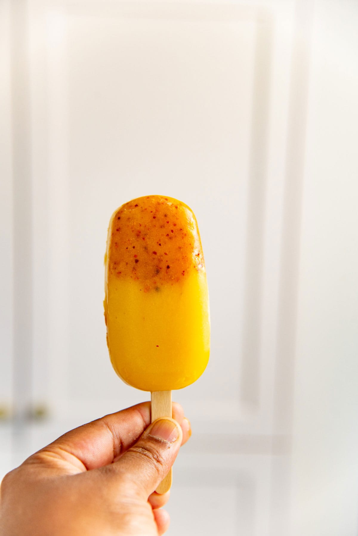 A hand holding up a mango tajin popsicles with a red spice layer at the top against a white background.