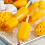 Mango popsicles on a bed of ice with fresh mangoes next to them. Some popsicles have a spicy tajin layer at the tip.