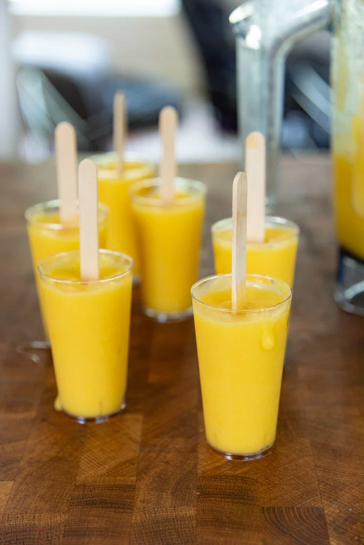 Small 3 oz disposable plastic cups filled with mango popsicle mixture and popsicle sticks put in place.