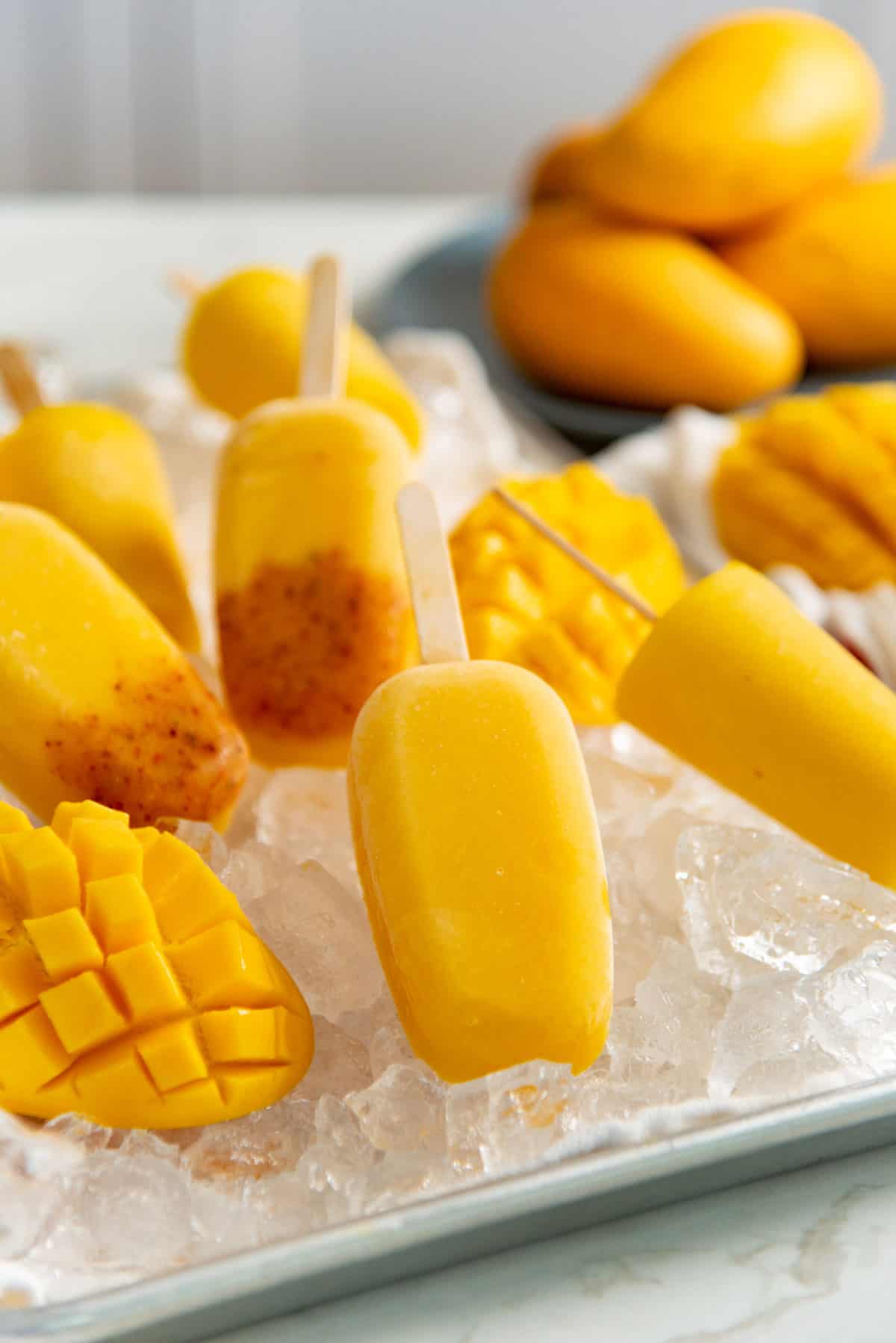 Bright yellow mango popsicles and mango tajin popsicles placed on top of ice. Fresh mangoes on a small plate in the background.