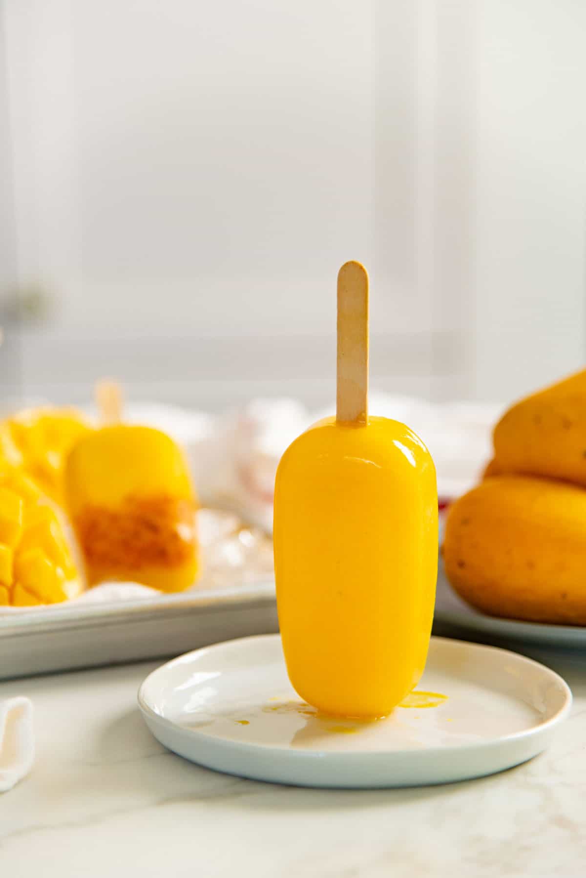 Bright yellow mango popsicle standing upside down on a small white plate, with more popsicles and fresh mangoes in the background.