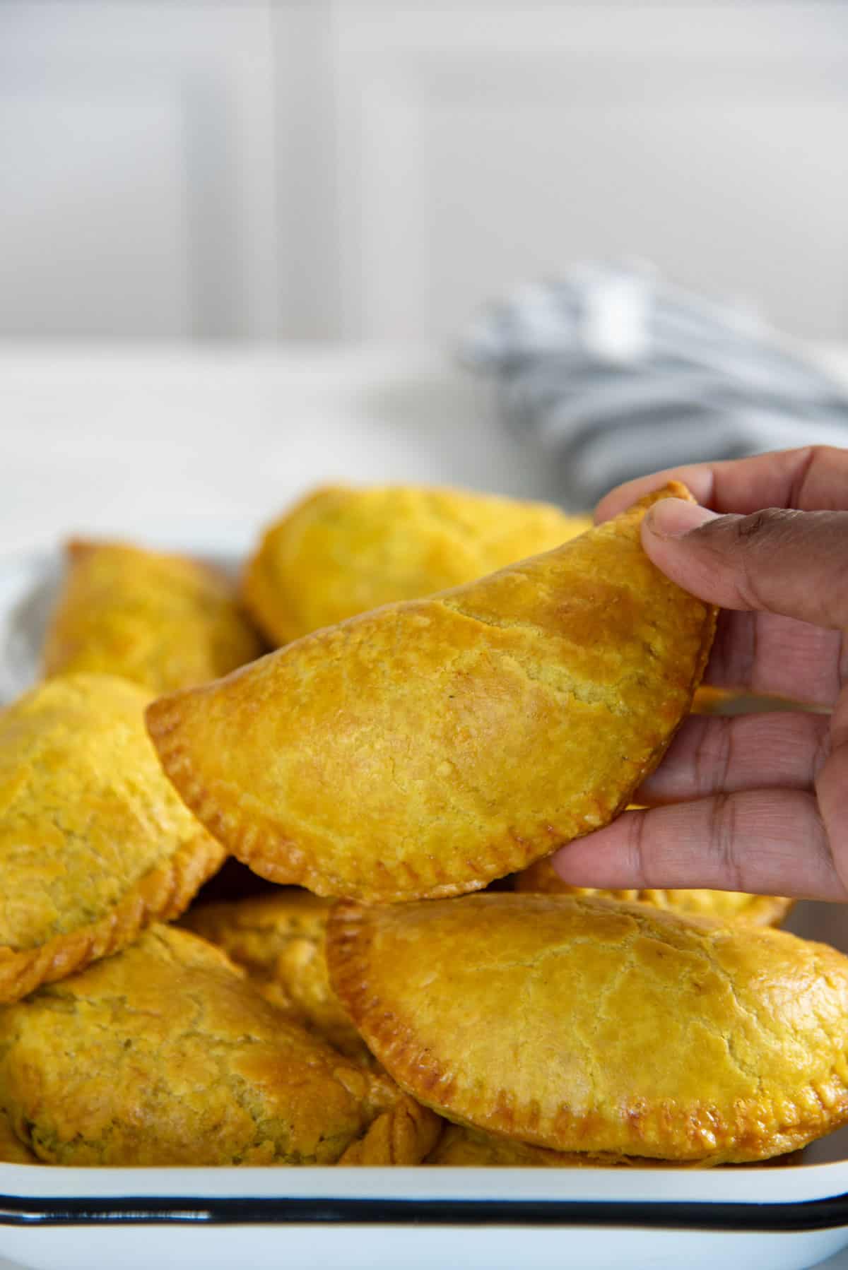 A hand holding a freshly baked golden Jamaican beef patty, with more baked patties in the background.