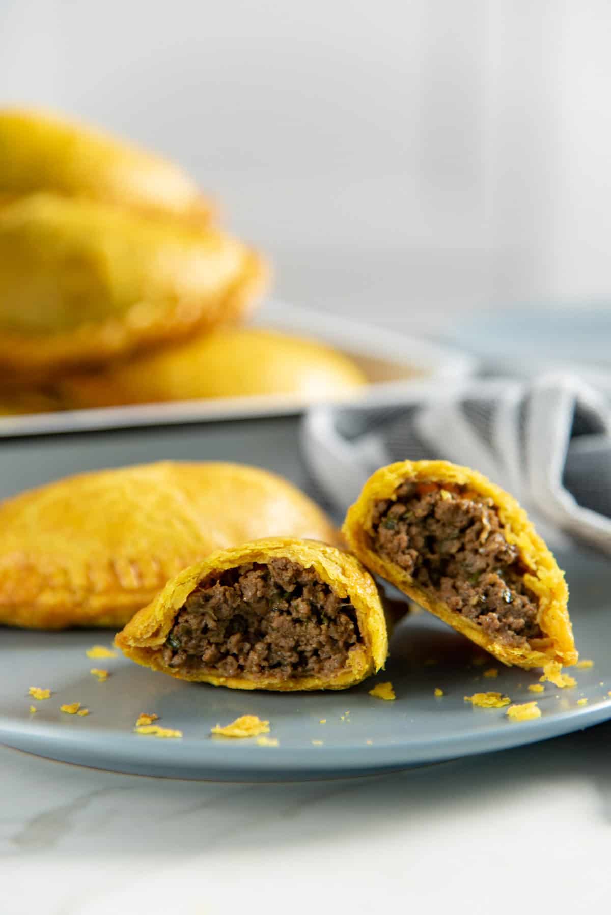 Close up of a beef patty broken in half to show the juicy, spicy ground beef filling in the middle. Flaky pastry crumbs are also seen next to it.
