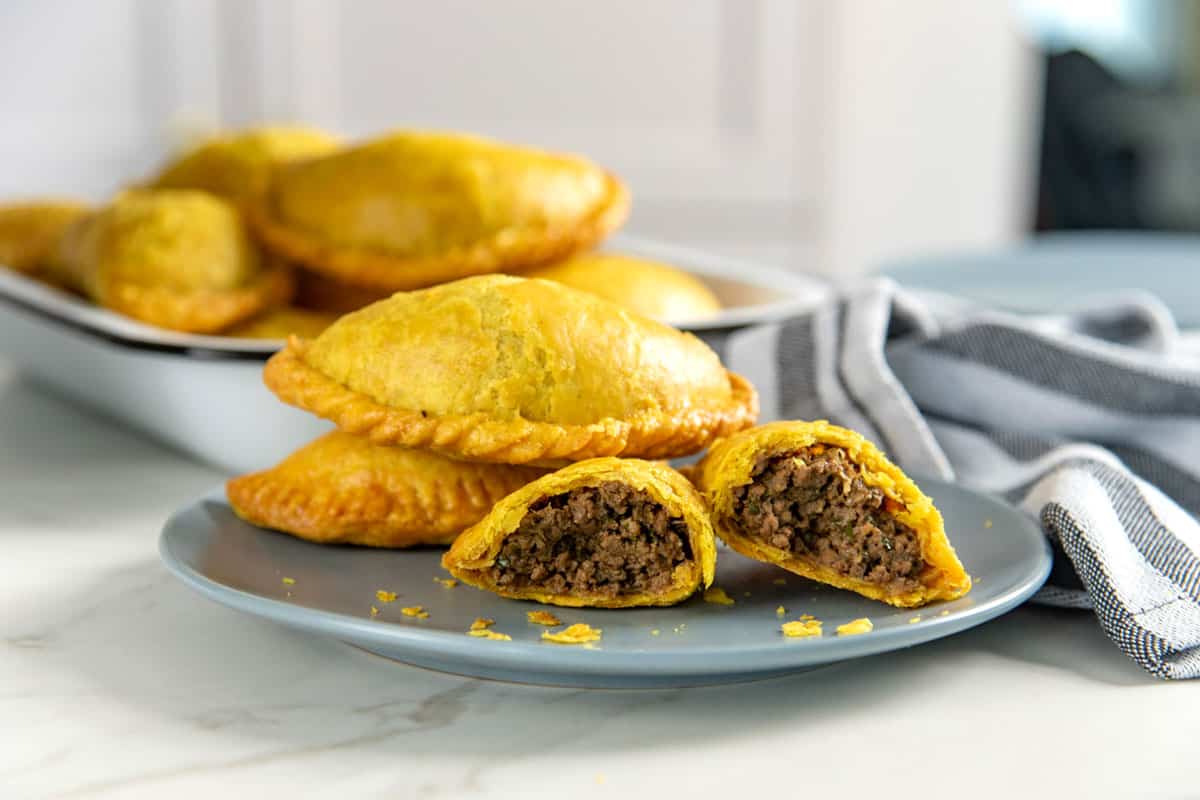 Three yellow, flaky Jamaican beef patties on a small blue plate, with one broken in half to show the filling inside.