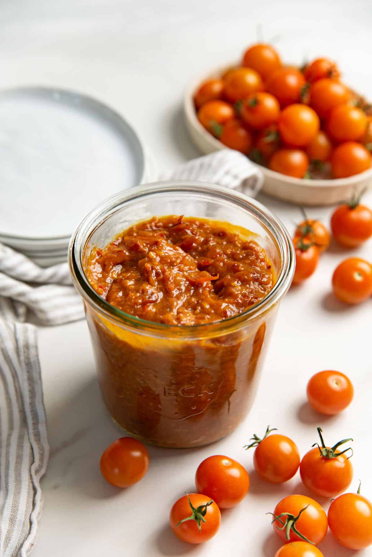 Jammy, chunky bacon tomato jam in a glass jar, with lots of fresh cherry tomatoes next to it on a white table.