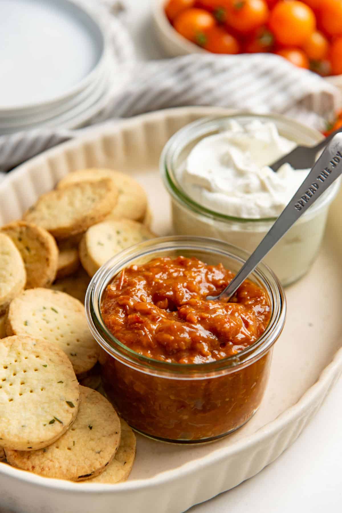 Chunky tomato jam in a glass jar with a spoon in it, with cheese crackers and sour cream on a platter next to it.