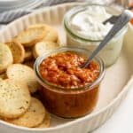 Dark reddish brown bacon tomato jam in a glass jar with a spoon in it, served on a dish with cheese cookies.