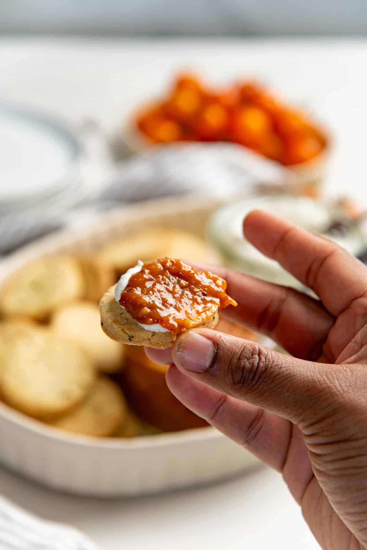A hand holding a cheese cracker topped with labneh and bacon tomato jam. More crackers can be seen in the background.