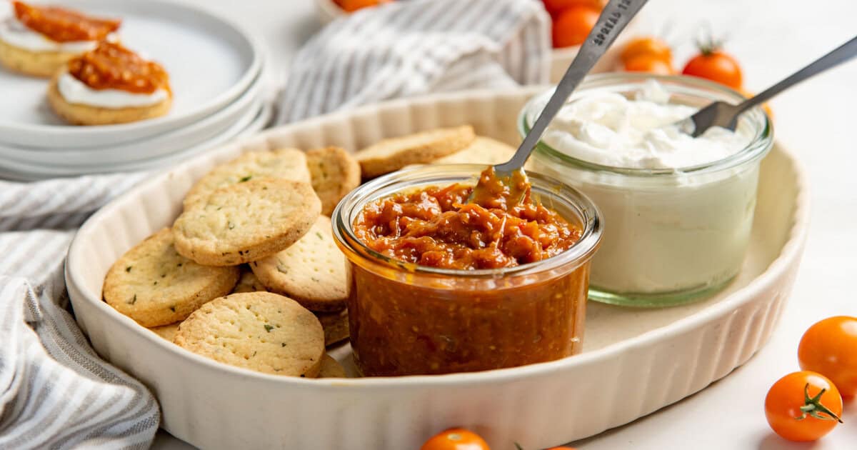 Reddish brown chunky bacon tomato jam served in a glass jar, along with savory cookies and labneh on a plate.