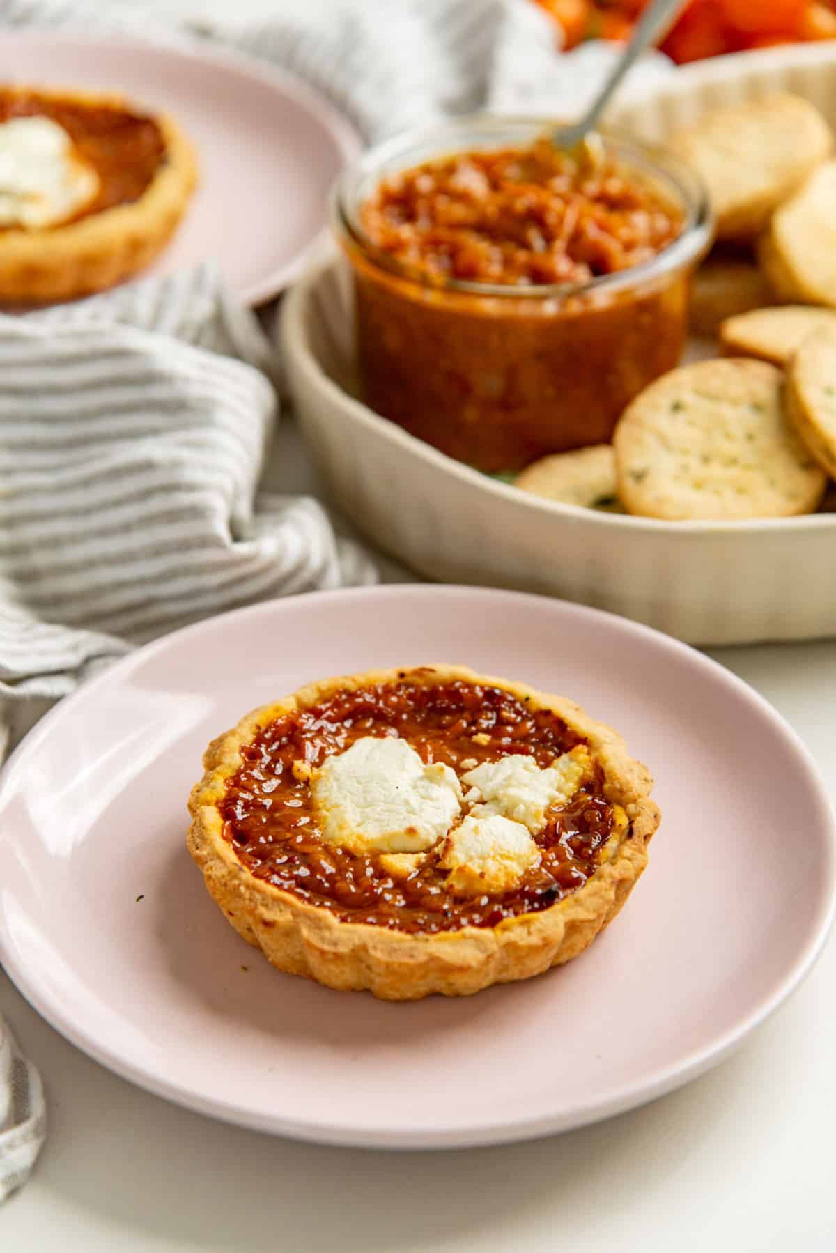 Savory tomato tart filled with bacon tomato jam and topped with feta cheese. A plate with cheese cookies in the background.