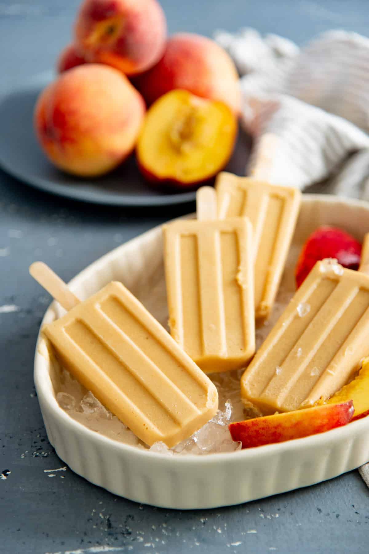 Several frozen peach popsicles on a bed of crushed ice in a dish, with fresh slices of peach next to them.
