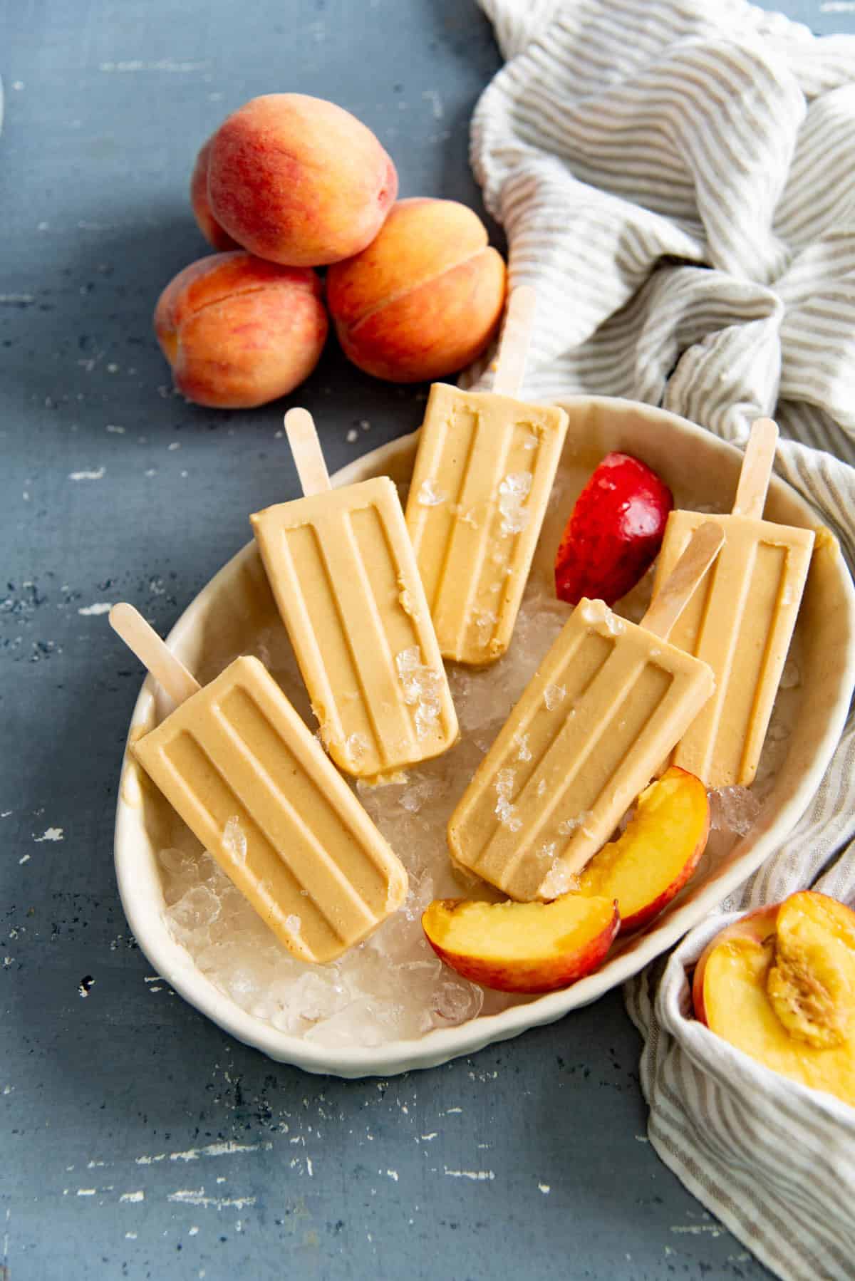 Several peaches and cream popsicles placed on a bed of ice with fresh peach slices next to them.