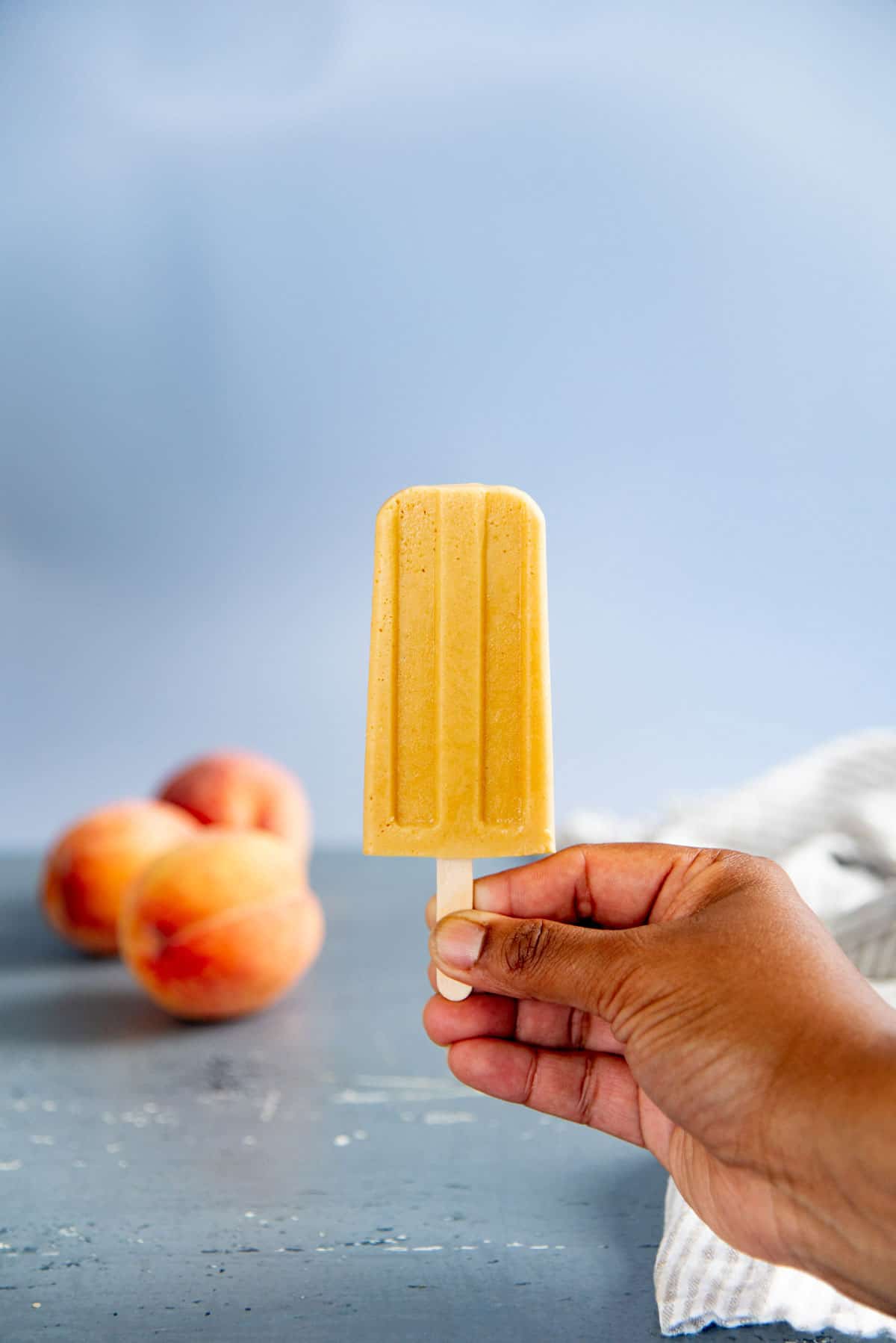 A hand holding a creamy popsicle by the popsicle stick. Several fresh peaches can be seen in the background.