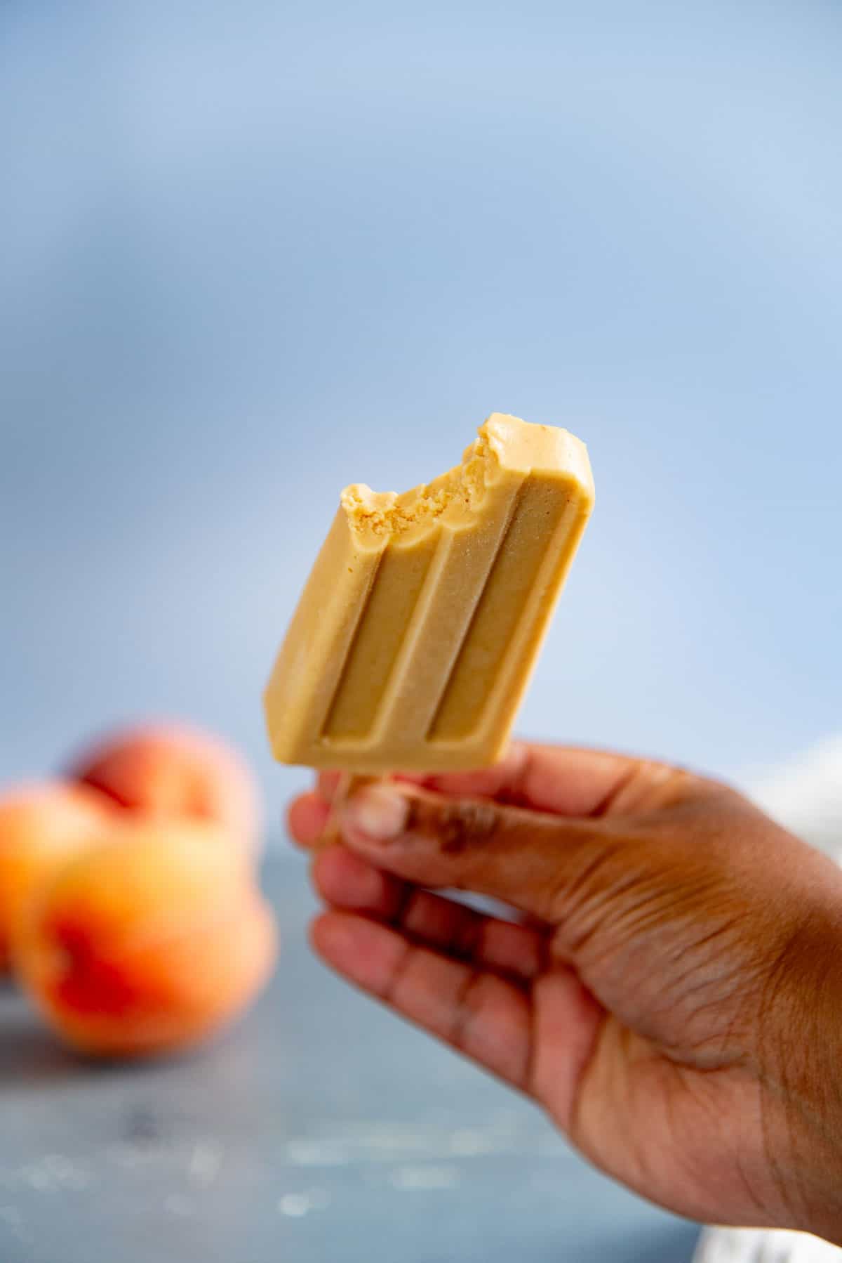 A bite taken out of a fresh peach popsicle, showing its creamy texture. Fresh peaches can be seen in the background.