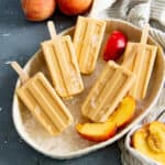 Overhead view of creamy peach popsicles in a dish with crushed ice, with fresh peaches and peach slices next to them.