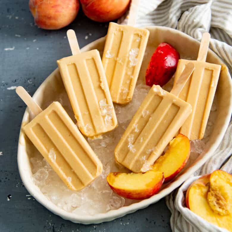 Overhead view of creamy peach popsicles in a dish with crushed ice, with fresh peaches and peach slices next to them.