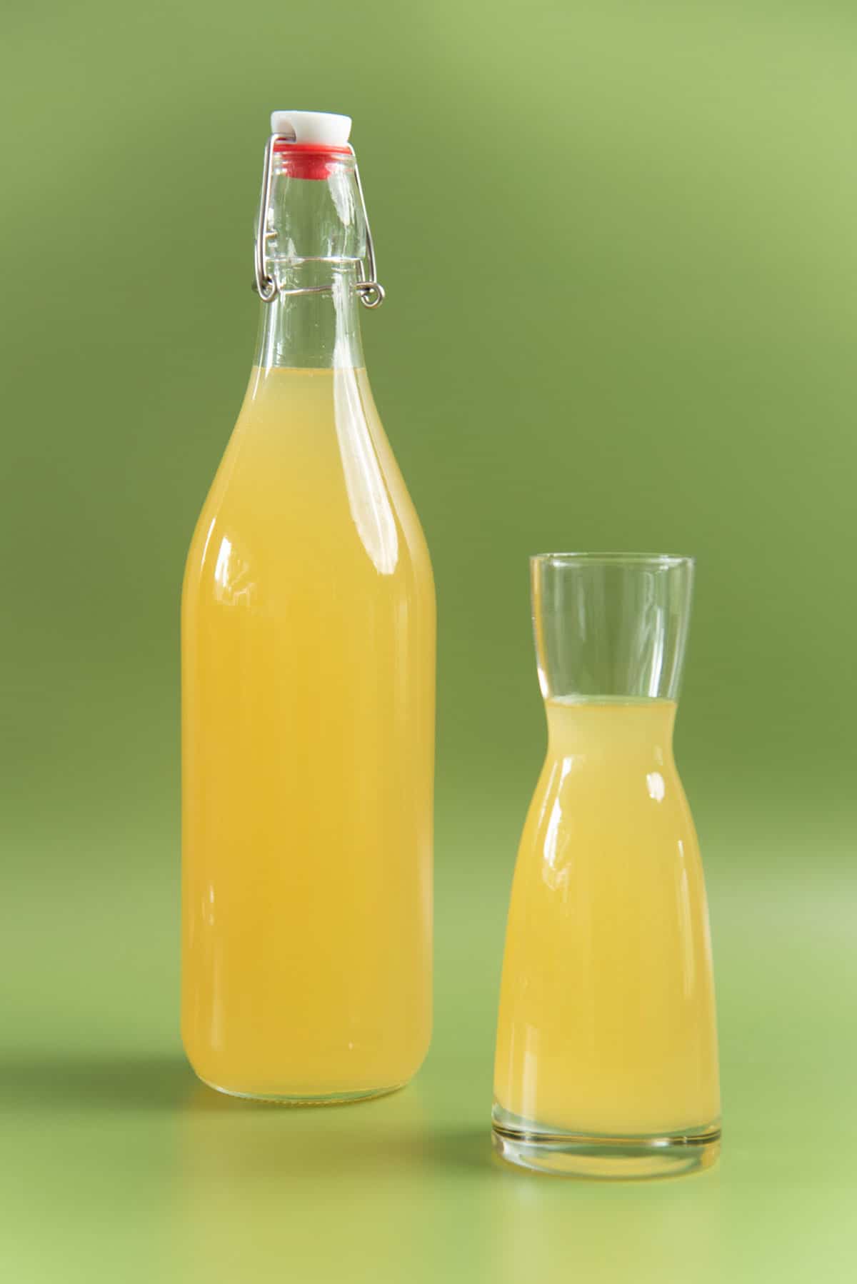 Elderflower cordial in a tall glass bottle and a short glass against a bright green background.
