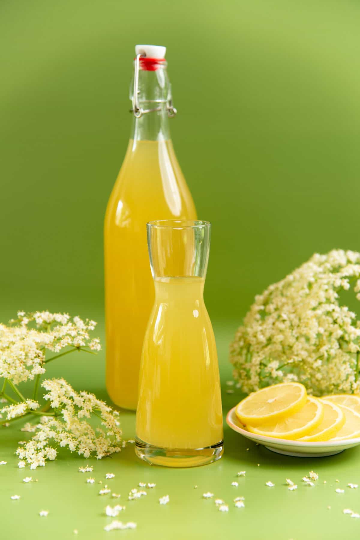 Elderflower cordial in a tall bottle and a short glass, and fresh elderflowers against a green background.
