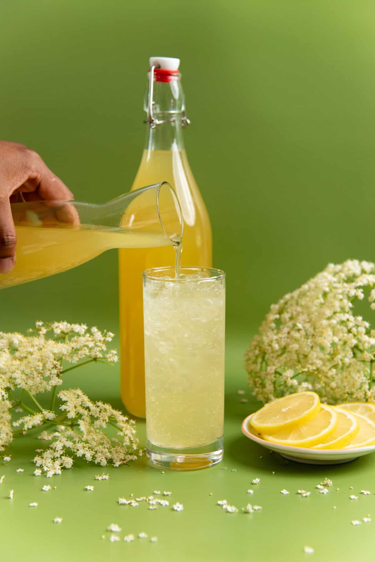 Pouring elderflower cordial into a tall glass with crushed ice. Lemon slices and fresh elderflowers are next to them.