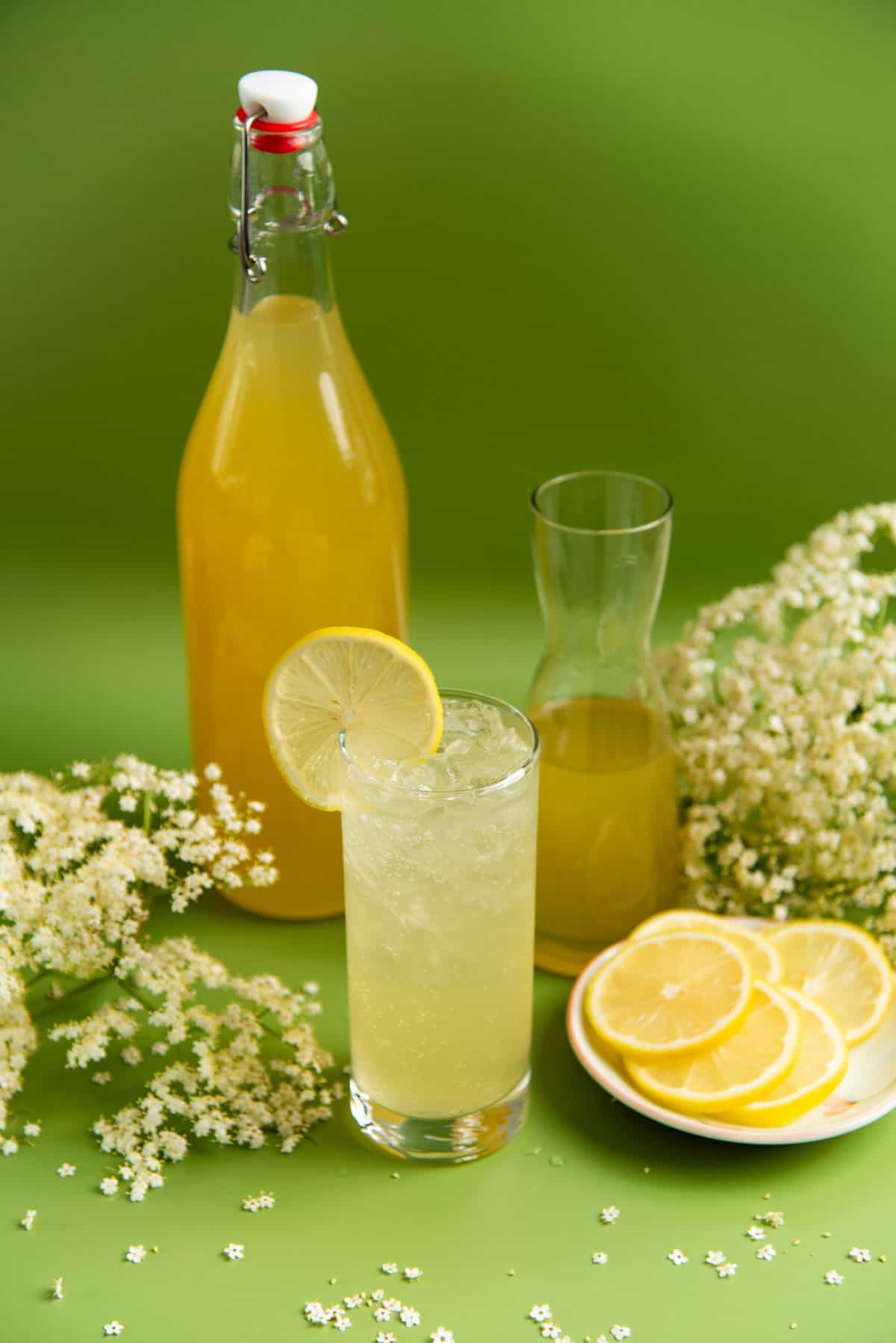 Another view of an elderflower lemonade in a glass, with the elderflower syrup in a tall bottle behind it.