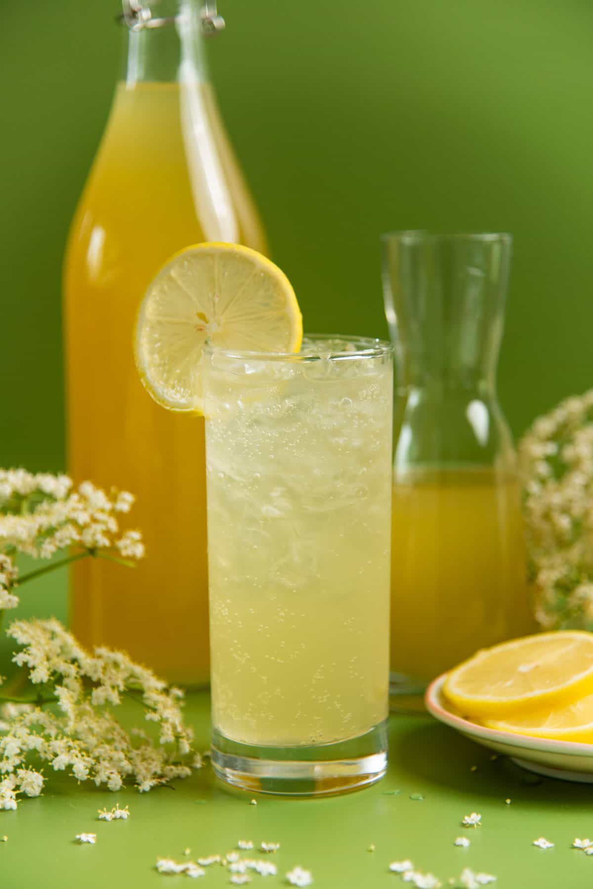 Close up of an elderflower lemonade in a glass, and elderflower cordial in a tall bottle behind it.